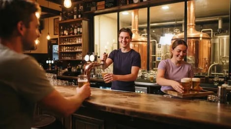 Barman tapt een speciaalbier achter de bar van een brouwcafé, terwijl een collega glazen bier op een plank zet, met koperen brouwketels achter glas op de achtergrond