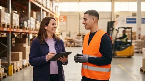 Intercedent met een tablet in gesprek met een uitzendkracht in een oranje veiligheidsvest in een magazijn, met een heftruck op de achtergrond